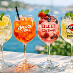 Vier erfrischende Spritz-Cocktails – Limoncello Spritz, Aperol Spritz, Lillet Wild Berry und Martini Lemon Spritz alkoholfrei – serviert in Weingläsern mit Früchten und Minze vor maritimer Kulisse